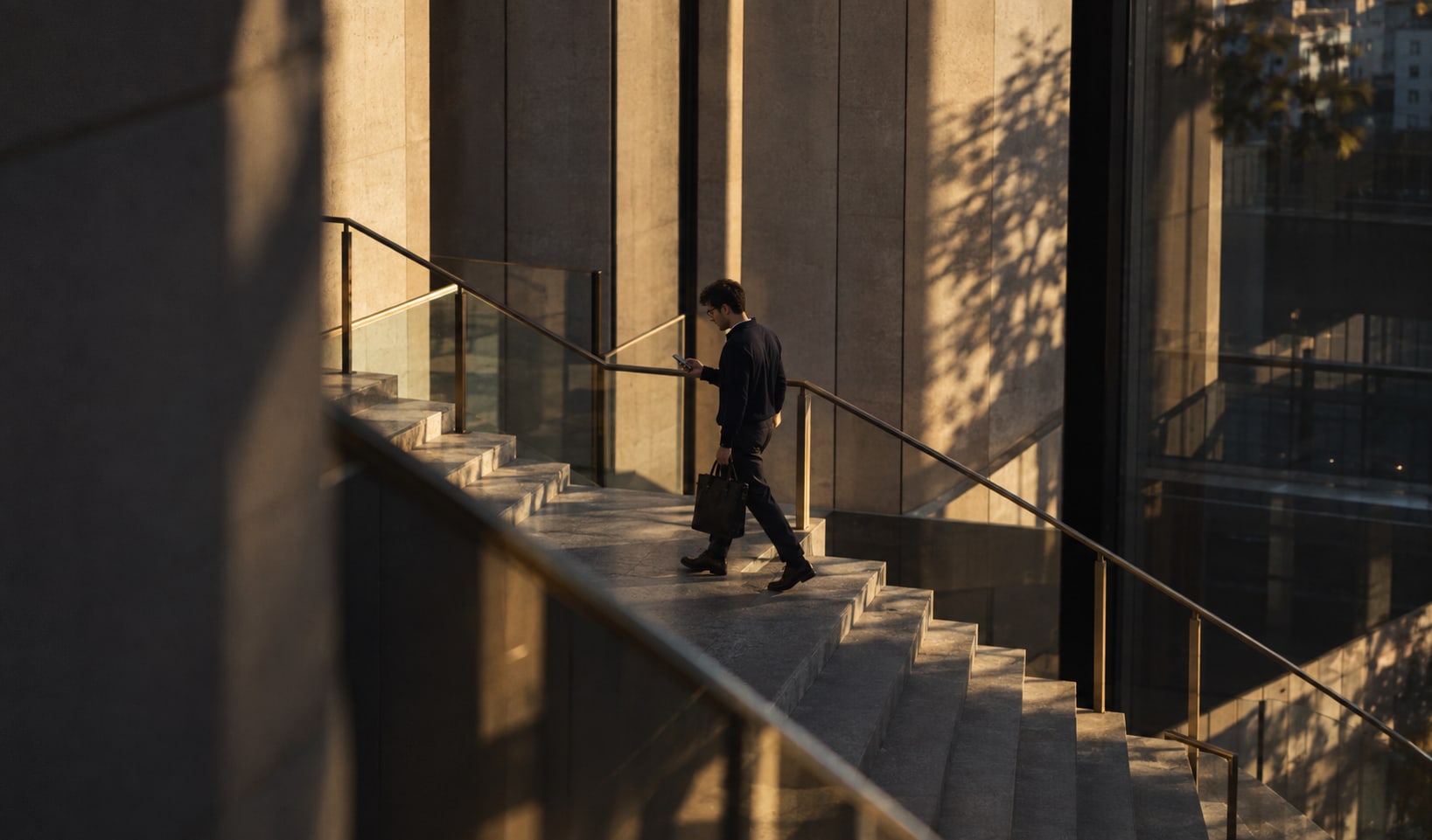 A professional with a briefcase walking up a sunlit stone staircase, checking his phone on the way to a meeting.